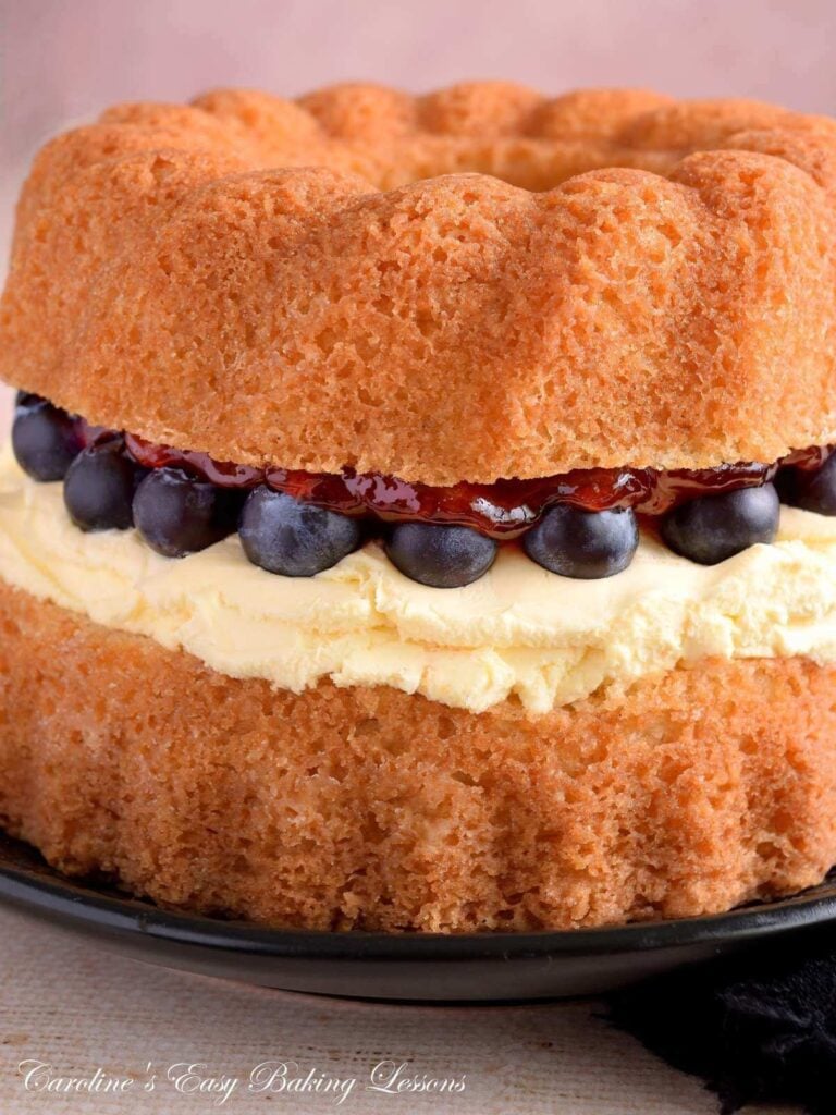 Extra close, partial vertical image of a gluten-free air fryer Victoria Sandwich bundt cake in layer form, showing the shinny strawberry jam, clotted cream and bluberries in detail.