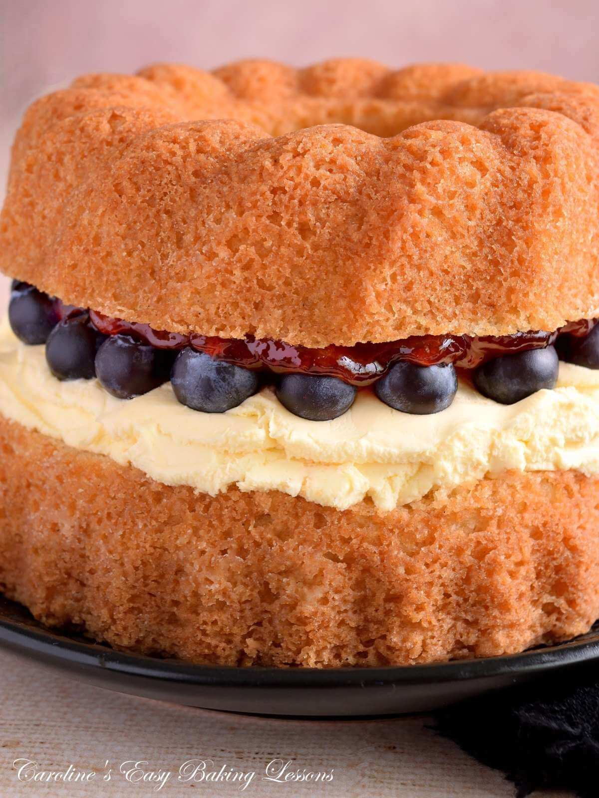Extra close, partial vertical image of a gluten-free air fryer Victoria Sandwich bundt cake in layer form, showing the shinny strawberry jam, clotted cream and bluberries in detail.