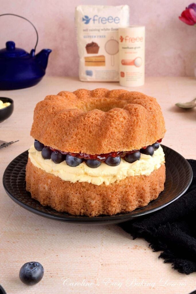 Close vertical shot of a table with black plate and teapot, large golden Victoria Sandwich bundt layer cake, showing clotted cream, jam and bluberry filling up close, and Freee Foods brand gluten free flour in the background.