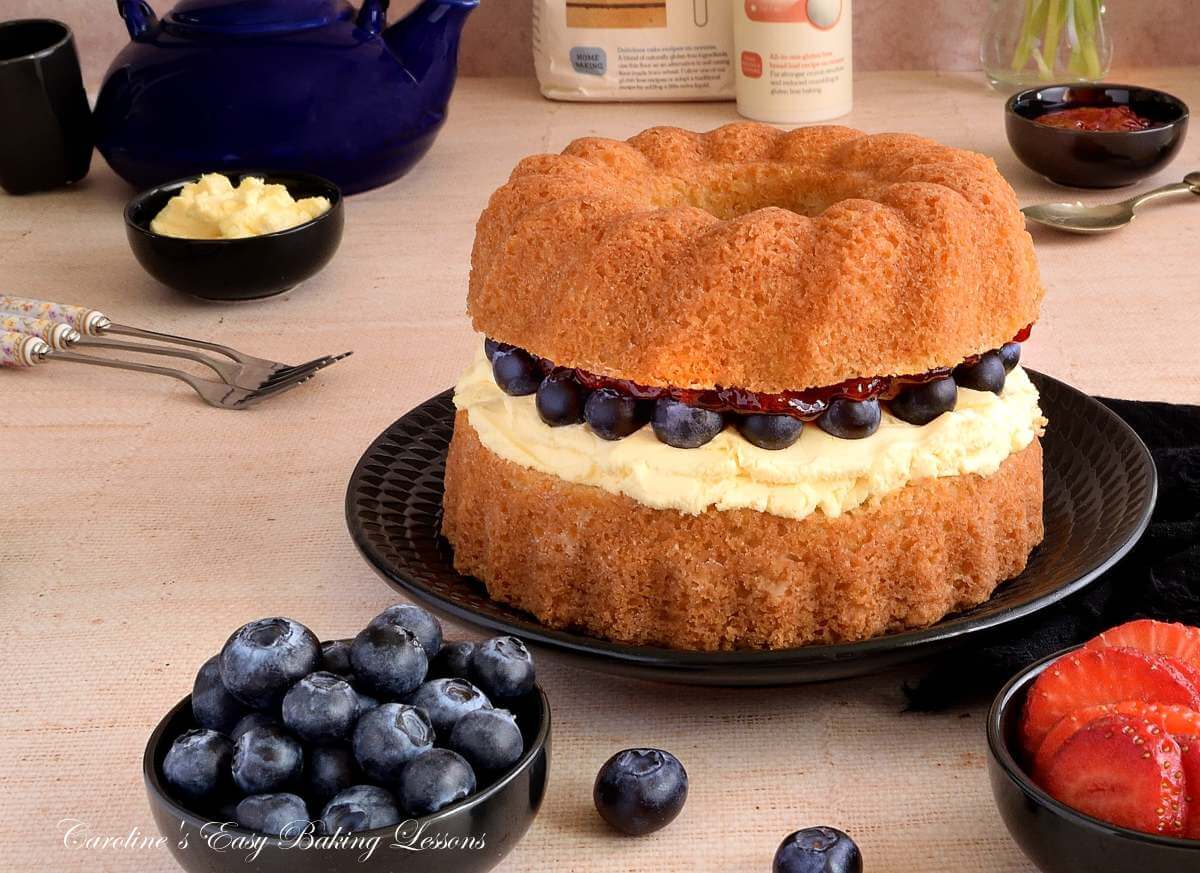 Horizontal colourful shot of a table with black crockery around a gluten-free air fryer Victoria sandwich bundt cake, layered with clotted cream, jam and blueberries, which also ar ein small pinch bowls surrounding the cake.