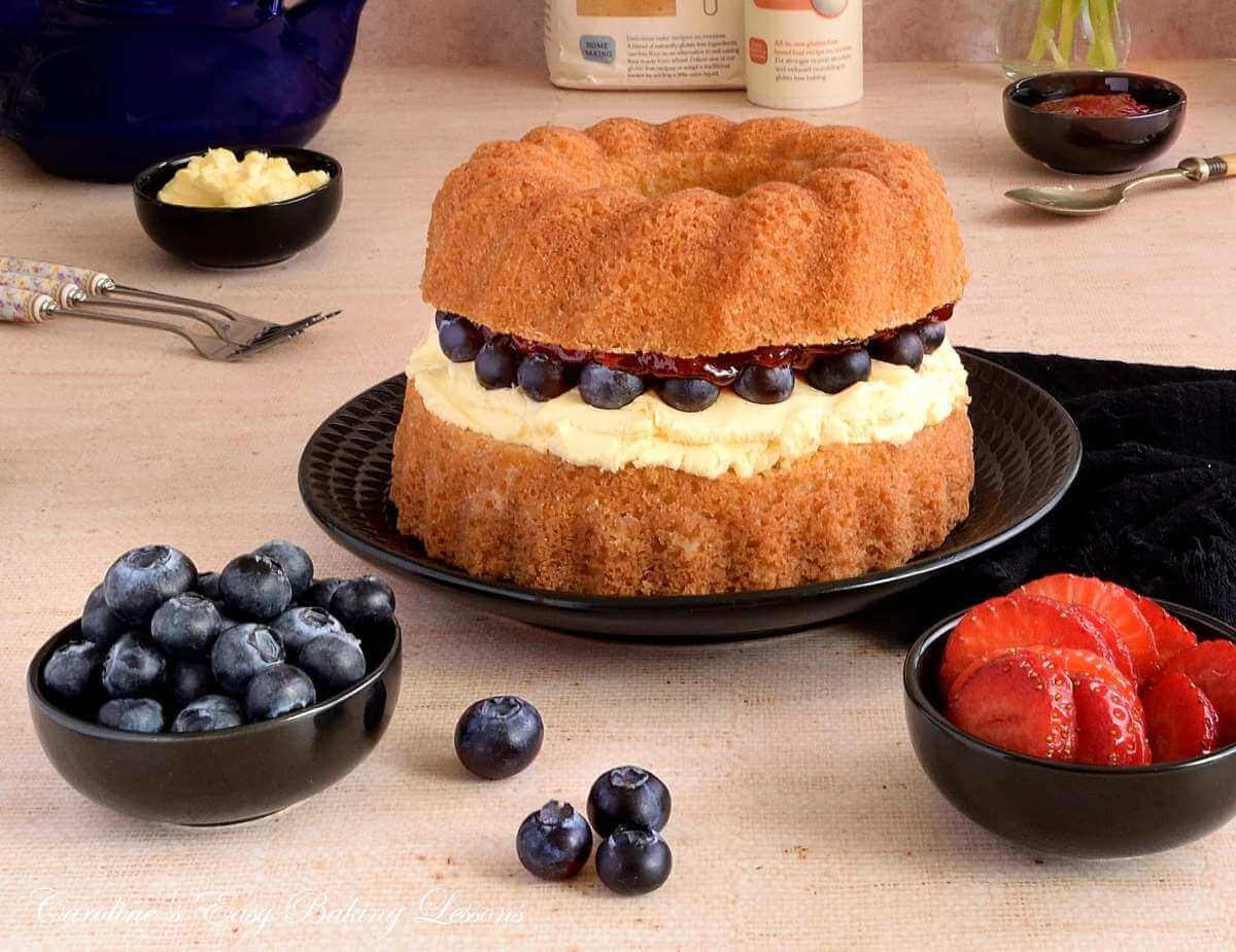 Horizontal image of talble with black and navy blue crockery around a golden Victoria Sandwich bundt cake, with clotted cream, jam and blueberry filling, and bowls of strawberry and blueberries to the side.