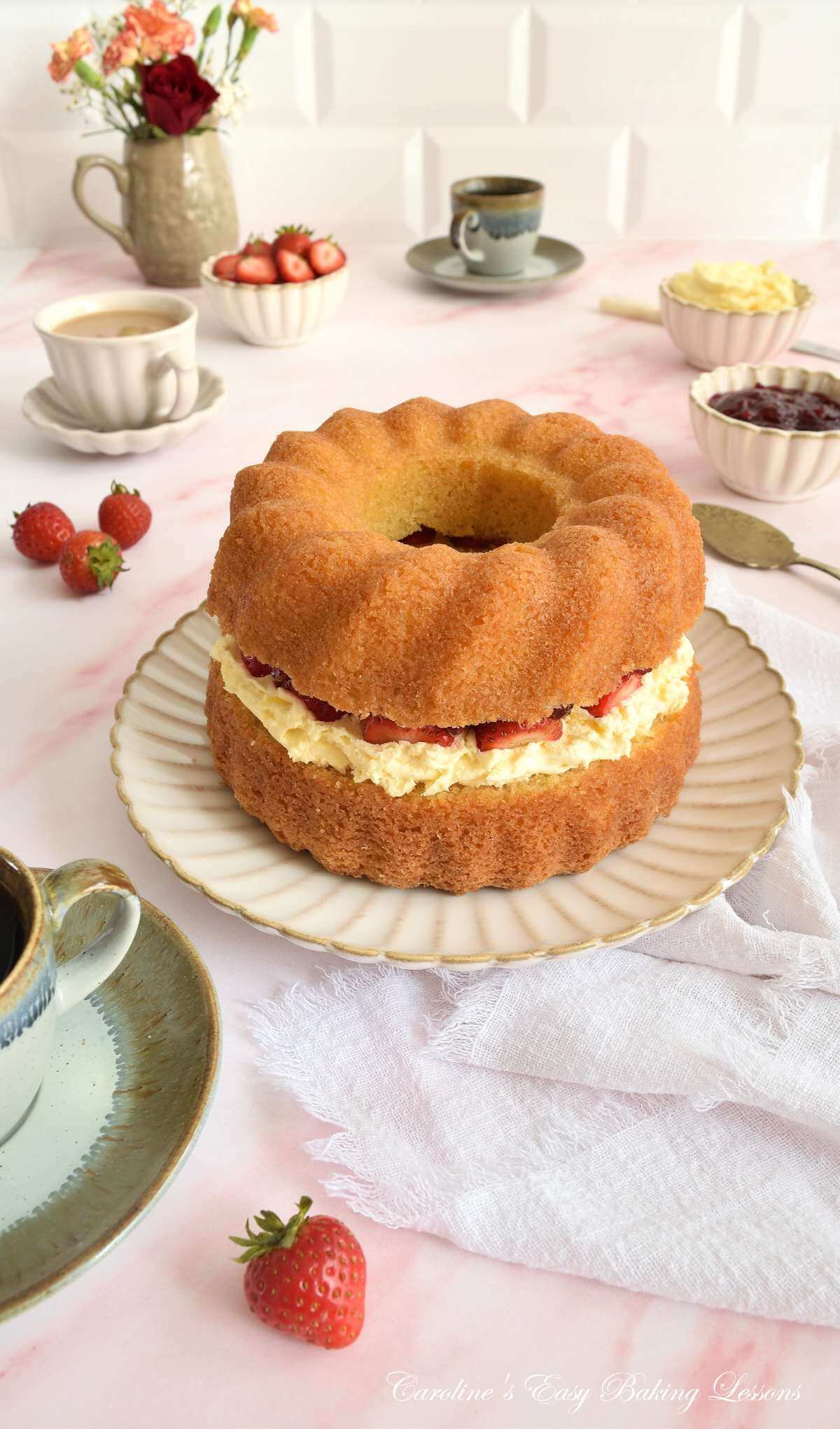 Long image of a pink marbled tabletop with beige crockery and linen, around a plate with a gluten-free Victoria Sandwich bundt cake, with a strawberry and clotted cream filling.