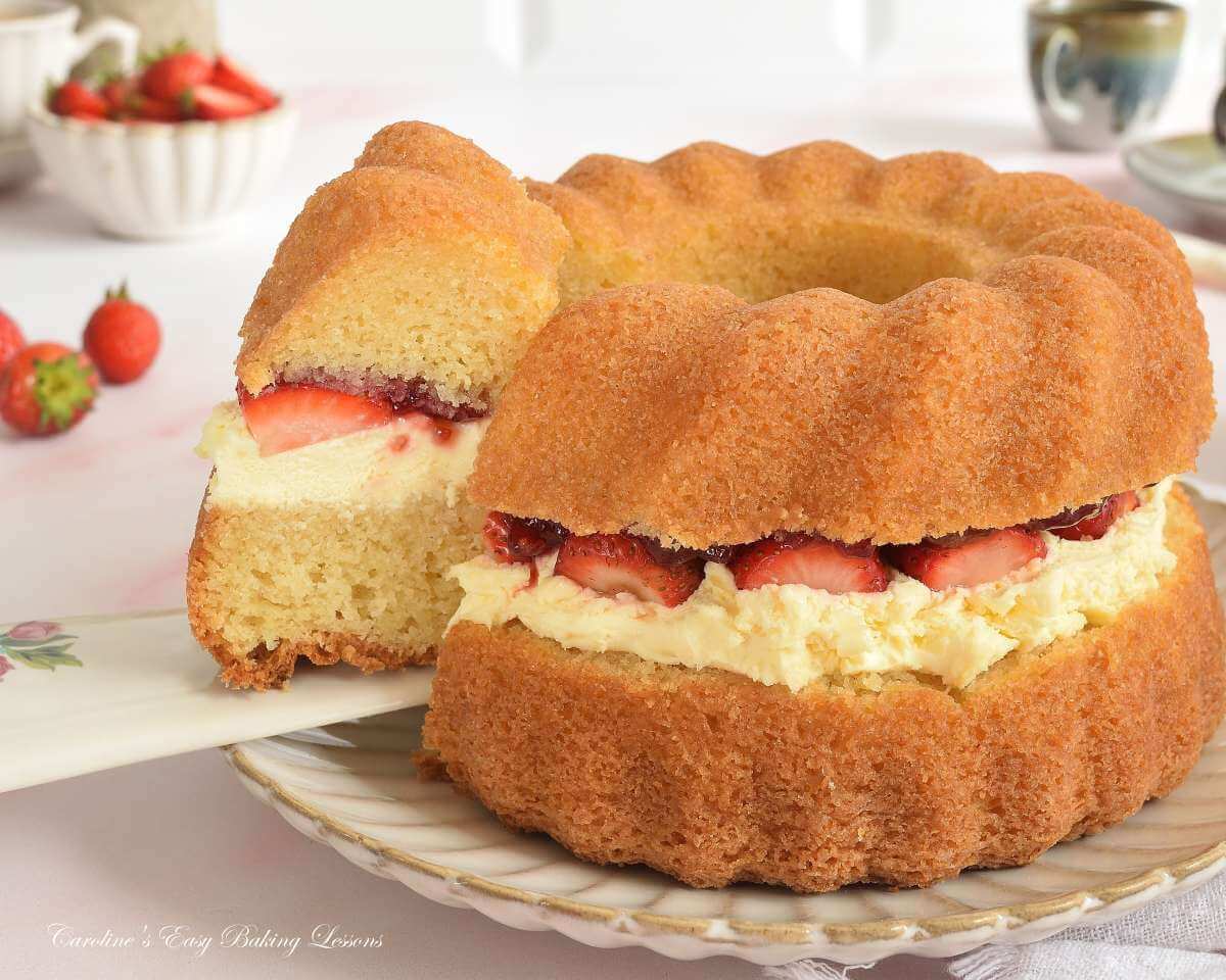 Close horizontal image of a golden Victoria Sandwich bundt cake, with cream and strawberry filling and a slice being removed.