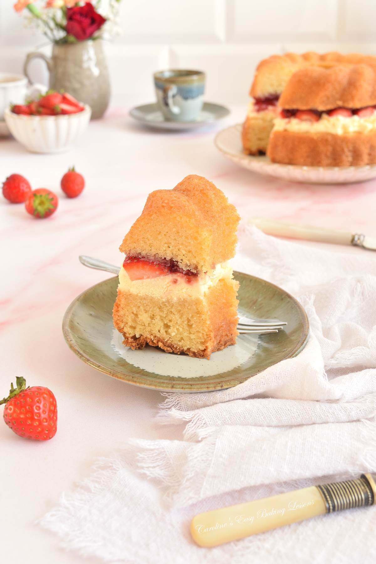 Vertical image of a light airy table with beige crockery, around a slice of gluten-free air fryer Victoria sandwich bundt layer cake, with a clotted cream, jam and strawberry filling.