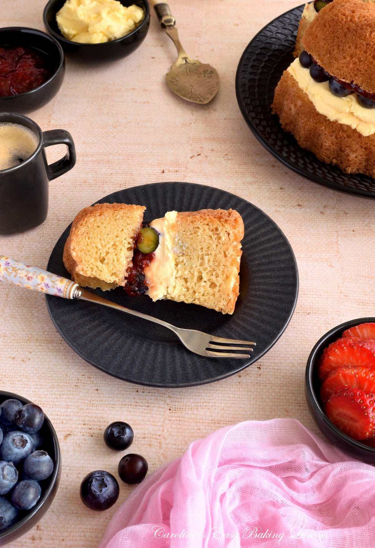 Close angled vertical shot of a table with a slice of Victoria Sandwich layered bundt cake, with cream, jam and bluberries, around pinch bowls of berries, coffee and the cake in the bakeground.