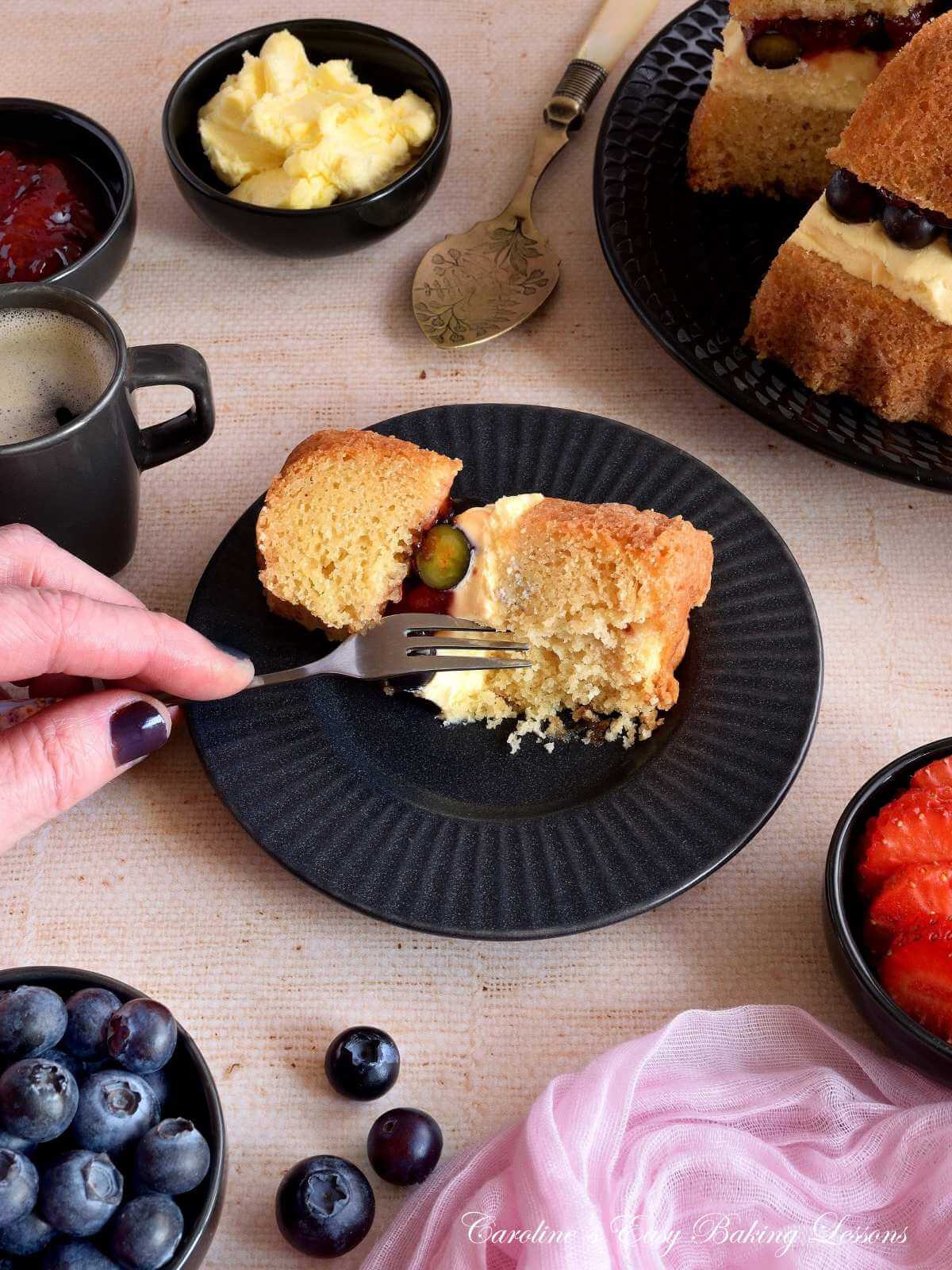 Overhead angled shot of a table with crockery and linen, pinch bowls of berries and a female hand cutting from a plate served with gluten-free Victoria sandwich bundt layer cake.