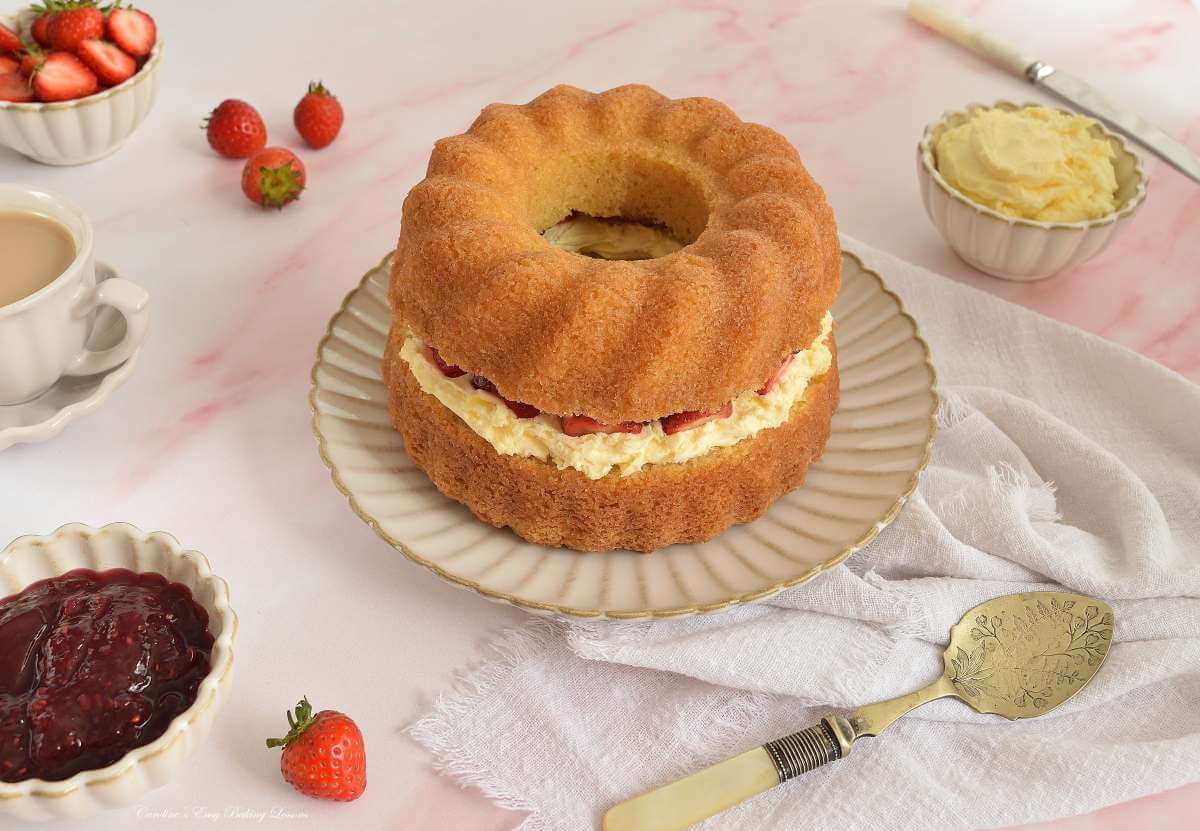Angled overhead image of a horizontal shot of pink marble table top with crockery around a golden Victoria sandwich layered bundt cake with cklotted cream and strawberries.