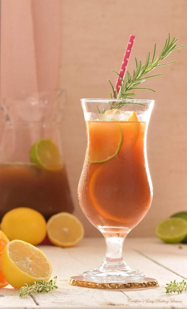 Vertical close image of a tall hurricane cocktail glass, with citrus iced tea, filled with ice, orange slice and rosemary garnish, straw and persparation on the glass, with citrus slices and pitcher in the background.