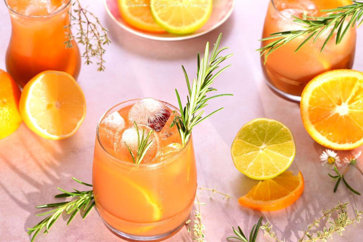 Horizontal image at 45 degrees of 3 bright citrus iced tea in glasses, with lots of ice, orange, lemon and lime slices, rosemary garnish and lots of sunny shadows.
