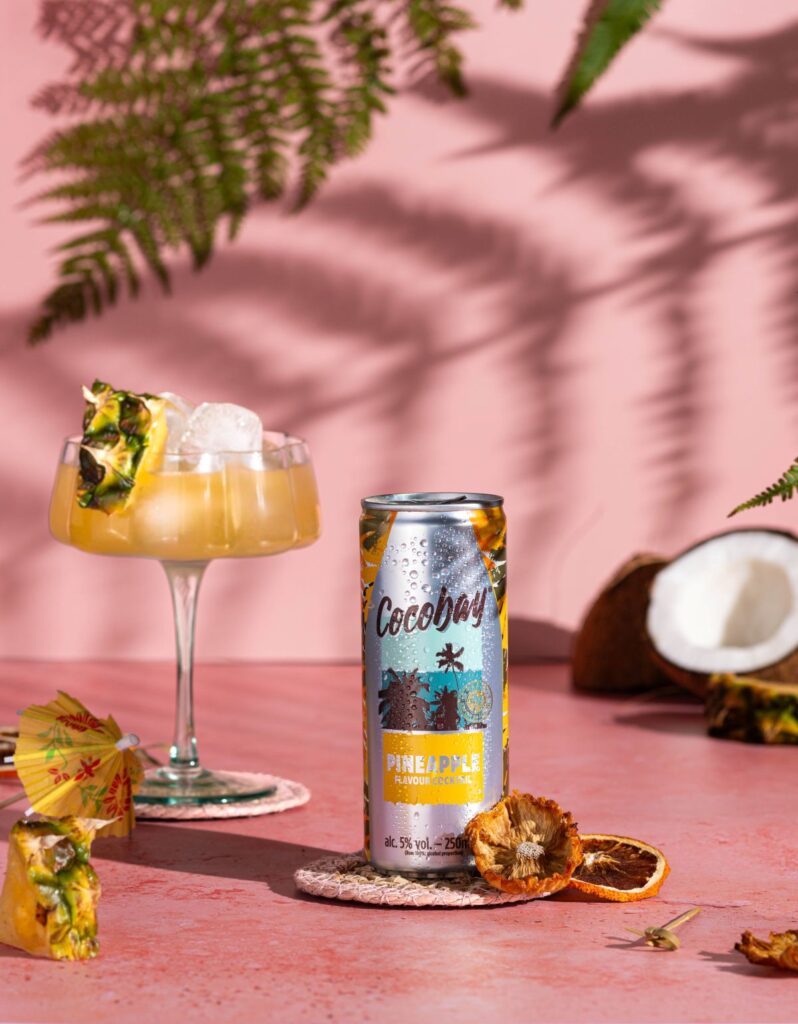 Bright image, on pink backdrops, with palm tree shadows, pineapple and coconut pieces, surrounding a glass and can of Aldi's pineapple cocktail drink.